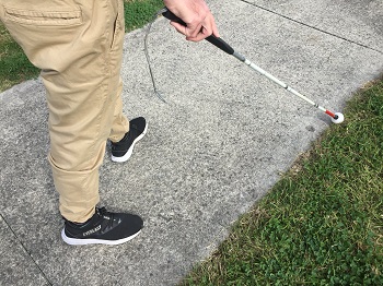 A close up image of a pair of male legs wearing beige coloured trousers, black runners and extending a white cane forward and to the right of the footpath against the edge of grass.