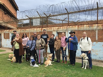 A group photograph of 12 people standing next to each other on grass, with a portion of the brick gaol wall seen in the background, including rusty spiralling barbed wire fencing extending beyond the brick fence above and behind our head height. Four dog guides and one Assistance Dog are also seen in close proximity to their handlers.