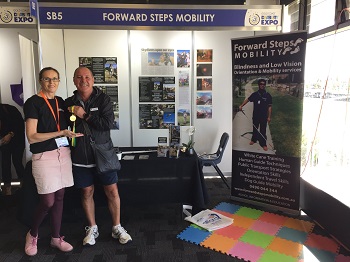 a photo of Kim and Tony standing side by side to left of Forward Steps Mobility booth with Tony showing his medal
