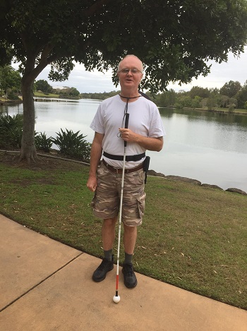 a photo of Peter holding a long white cane vertically in front of his body with grass and water behind