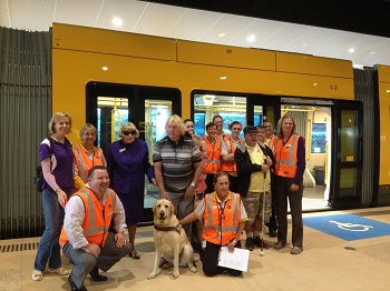 Image shows large guide dog Wesley centre of light rail platform with handler Kevin and surrounded by other people with tram in background