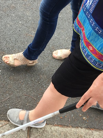 Two legs visible assuming a sighted guide technique with partial view of a long cane to the left