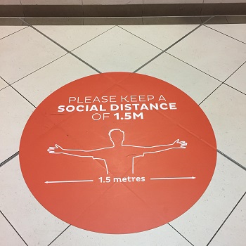 Photo shows a large round orange sign stuck to tiles on the ground stating, Please keep a social distance of 1.5 metres, and also includes a picture of someone stretching their arms out bilaterally