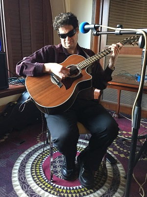 A terrific photograph of Steve sitting on a stool with microphone positioned in front of him on a stand, guitar in hand, and sunshine aptly beaming through a nearby window as though placing him in a naturally lit spotlight for his captive audience.