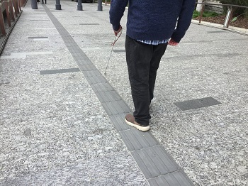 Photo taken from behind and to the left side of a male who is seen walking along a set of TGSIs in King George Square, with white cane extended forward. The image also shows the tiles extending further forward and off into the distance ahead.