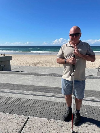 A picture of Dan standing on black tactile ground surface indicators near the top of some descending steps leading down onto the beach at Surfers Paradise. Dan is facing away from the beach with his long cane in vertical placement. The blue ocean, white sand and blue sky are seen in the background.