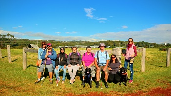 Image shows Kerri standing, with Justin, Kushal, Hailey, Jenny, Peter and Tarhnee all sitting side by side on a wooden gratitude seat at 'The Farm', near Byron Bay. Kim is standing far right of the group, with two black dog guides present